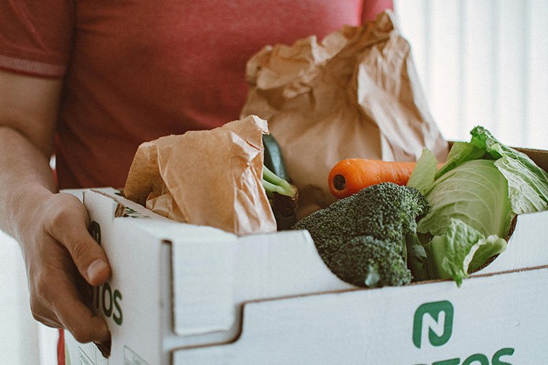 A volunteer holds a white box filled with fresh vegetables and bagged food items, ready for distribution to those in need.